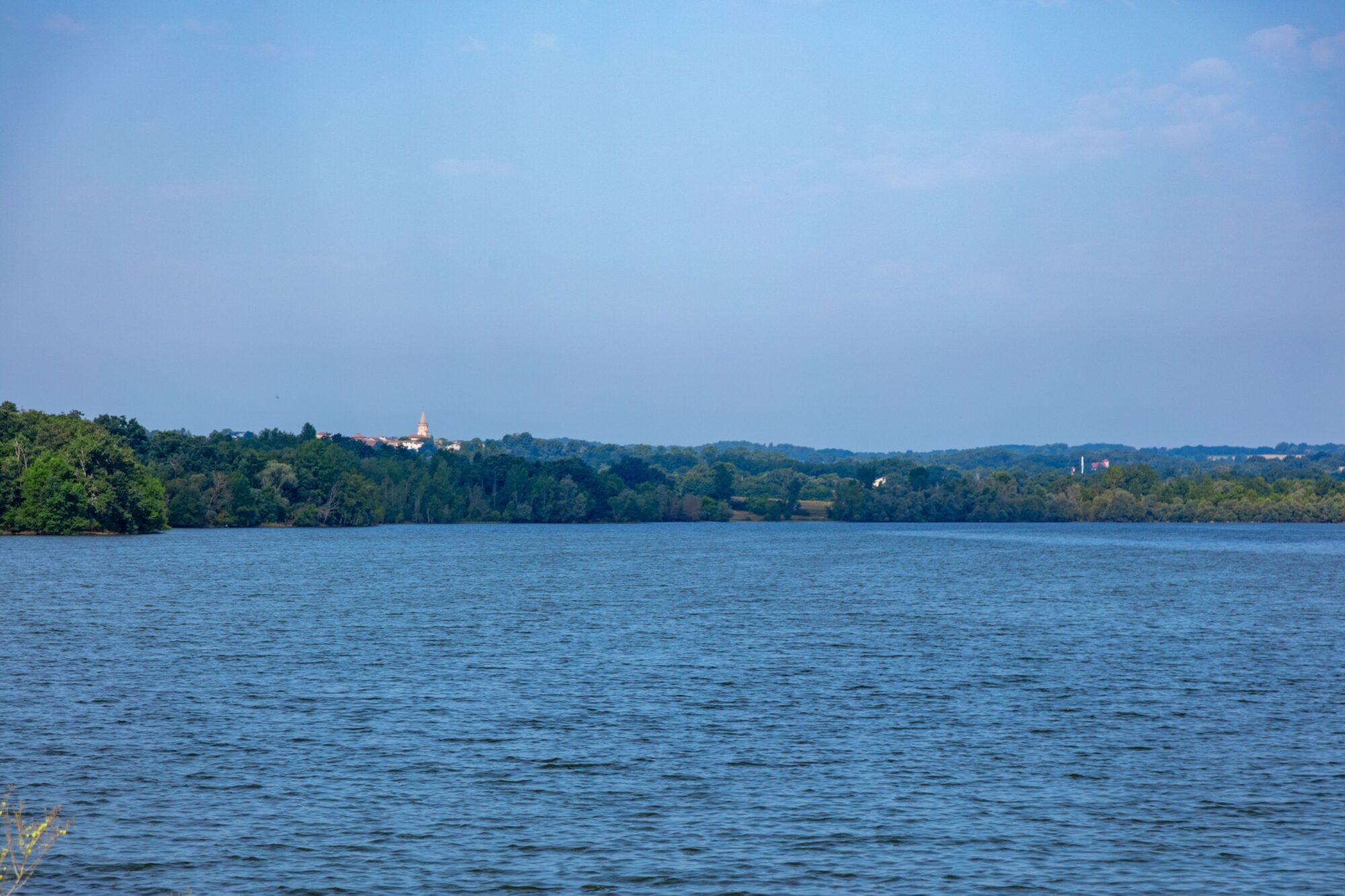 Lac bordé d’arbres avec clocher au loin, Office de Tourisme Cœur de Garonne
